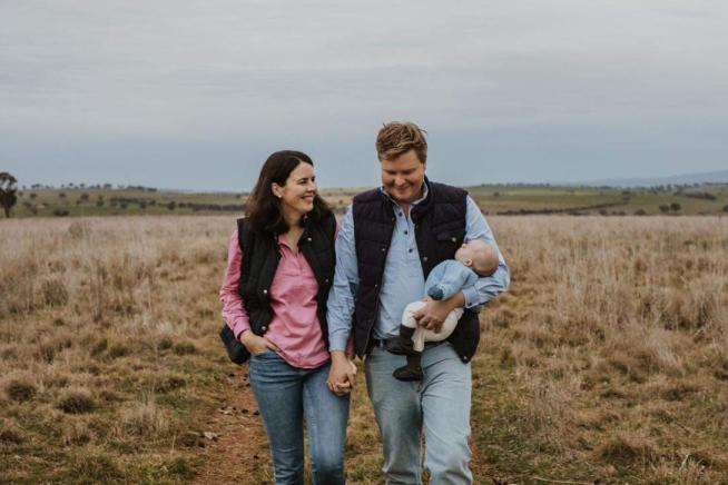 Woman and man holding baby, walking on farm paddock.